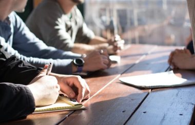 A number of people are shown sitting around the table. None of their faces are visible, you mostly see their hands - one of which is tapping the table as if making a point. The other people are leaning forward, their hands clasped and siting on the table