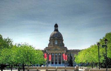 Vue sur l'Assemblée législative de l'Alberta. Il s'agit d'un bâtiment brun avec un toit en forme de dôme. Des arbres se trouvent à la gauche et à la droite de l'entrée.