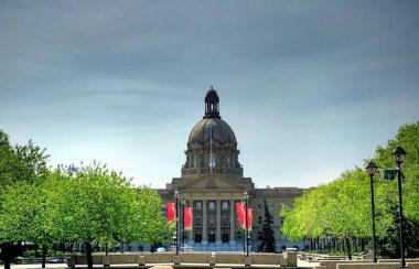 Vue sur l'Assemblée législative de l'Alberta. On aperçoit l'entrée de l'Assemblée. Le toit du centre du bâtiment est en forme de dôme. La couleur de la bâtisse est brune. Des arbres se retrouvent sur les côtés de l'entrée. Au centre de l'entrée, on voit des affiche rouge accroché sur des lampadaires.
