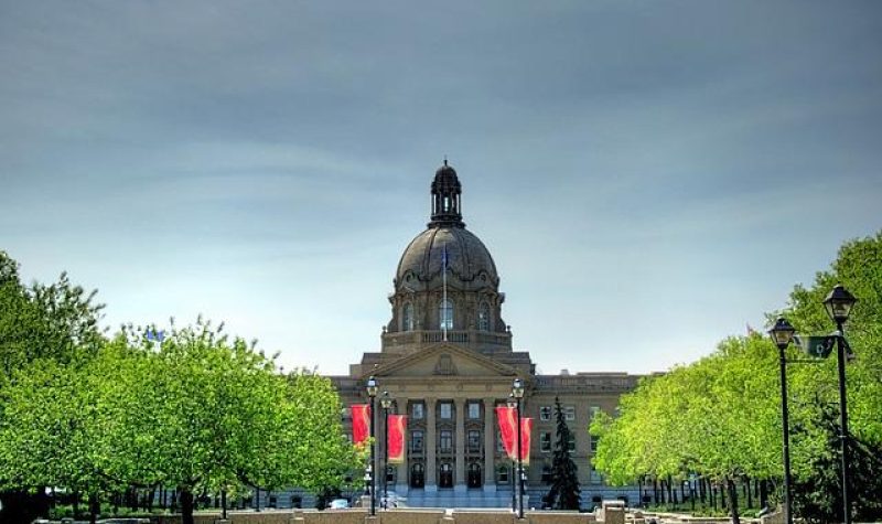 Vue sur l'Assemblée législative de l'Alberta. On aperçoit l'entrée de l'Assemblée. Le toit du centre du bâtiment est en forme de dôme. La couleur de la bâtisse est brune. Des arbres se retrouvent sur les côtés de l'entrée. Au centre de l'entrée, on voit des affiche rouge accroché sur des lampadaires.