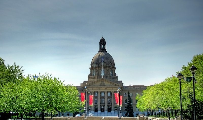 L'Assemblée législative de l'Alberta vu d'en face. On y aperçoit des arbres et des lampadaires devant. Des bannières rouges sont attachées aux lampadaires.