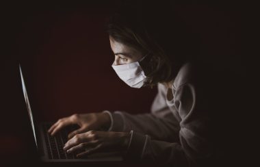 Une femme portant un masque blanc et un coton ouaté gris tape sur un clavier devant son ordinateur