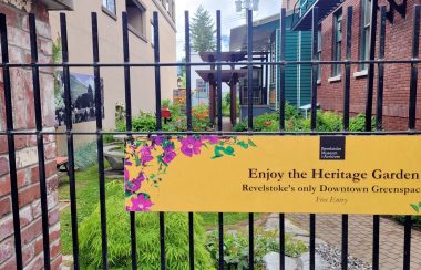 Yellow sign on an iron fence says, Enjoy the Heritage Garden, Revelstoke's only downtown green space. Brick wall surrounding a garden beyond the fence.