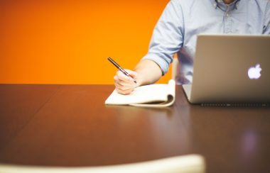 Un homme devant un ordinateur s'apprête à écrire. Il est assis sur une table en bois devant un fond orange.