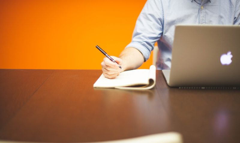 Un homme devant un ordinateur s'apprête à écrire. Il est assis sur une table en bois devant un fond orange.