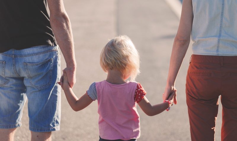 Two adults hold the hands of a small child who is walking between them. The child has blonde hair and a pink shirt. The adults are casually dressed and the photo is cropped below the shoulders of the adults.