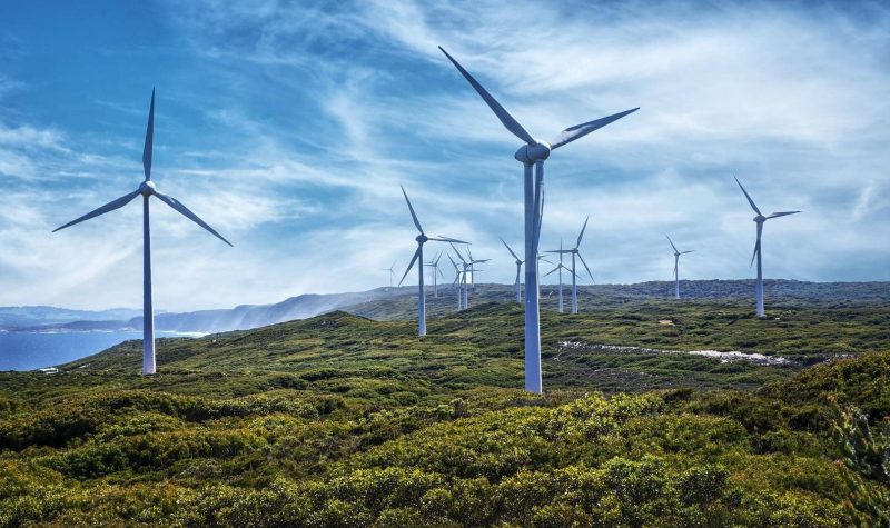 A number of tall wind mills stand across a green filed. The sky is blue and cloudy.