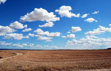 Un grand champ avec une route de campagne sur la gauche et un ciel bleu avec des nuages à perte de vue.