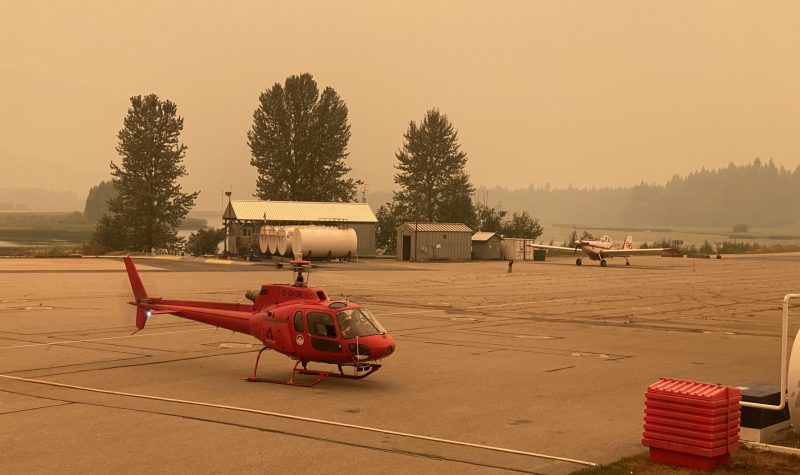 Red helicopter grounded on asphalt, smoky skies.