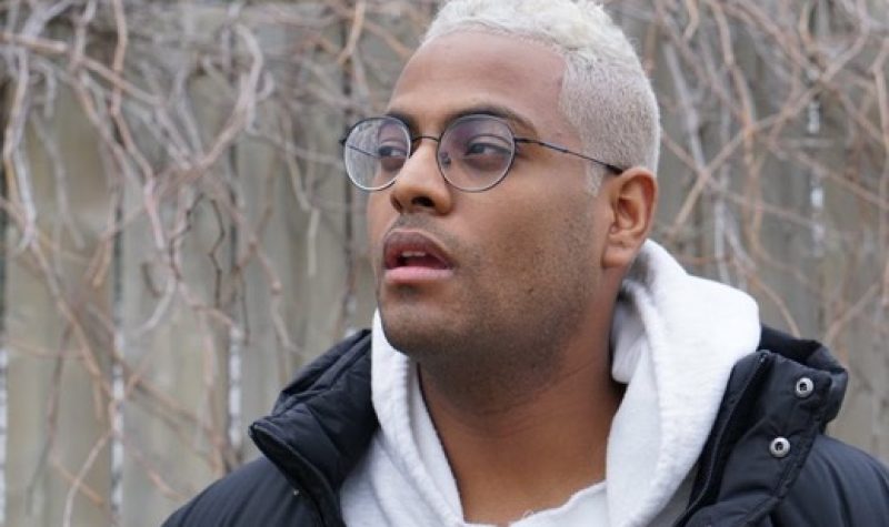 A headshot of Fitsum Arguy, with short white hair, a shite hoodie, a black jacket, and glasses, framed by naked trees in the background, and looking triumphantly towards the future.