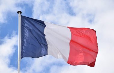 Le drapeau bleu blanc rouge de la France flotte dans le vent devant un ciel bleu et quelques nuages blancs.