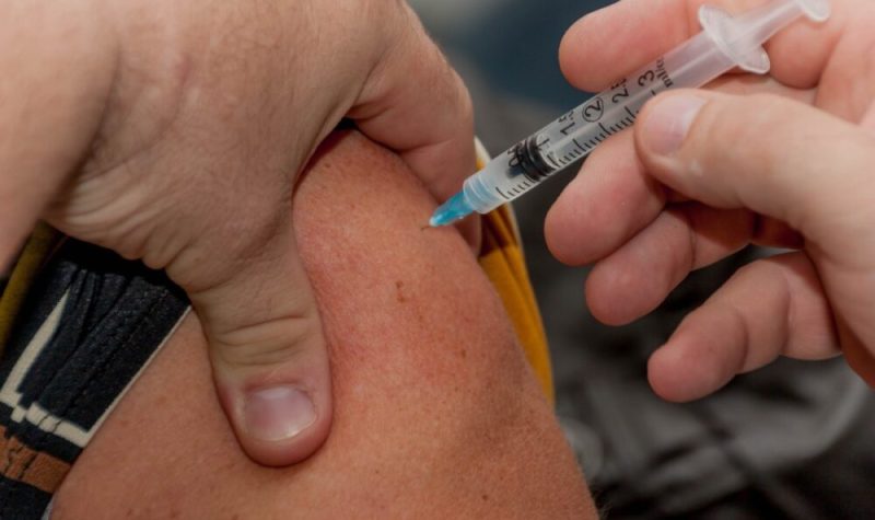 Two hands hold an upper arm as a white and blue needle with a flu shot is injected into the skin.