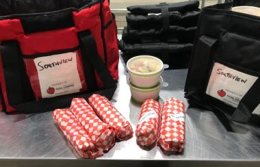 Two cooler bags are on a table, with five sandwhich wraps and two containers of food on the table between the bags.