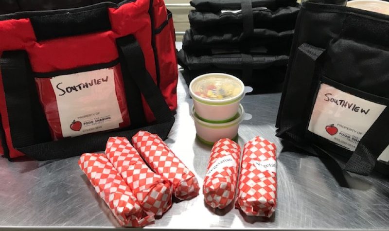 Two cooler bags are on a table, with five sandwhich wraps and two containers of food on the table between the bags.