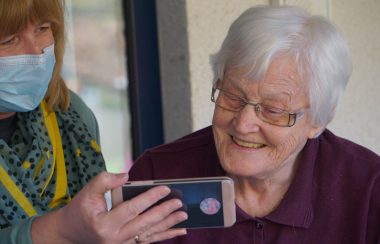 A younger woman with a face mask shows an elderly woman something on a smartphone.