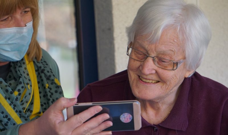 A younger woman with a face mask shows an elderly woman something on a smartphone.