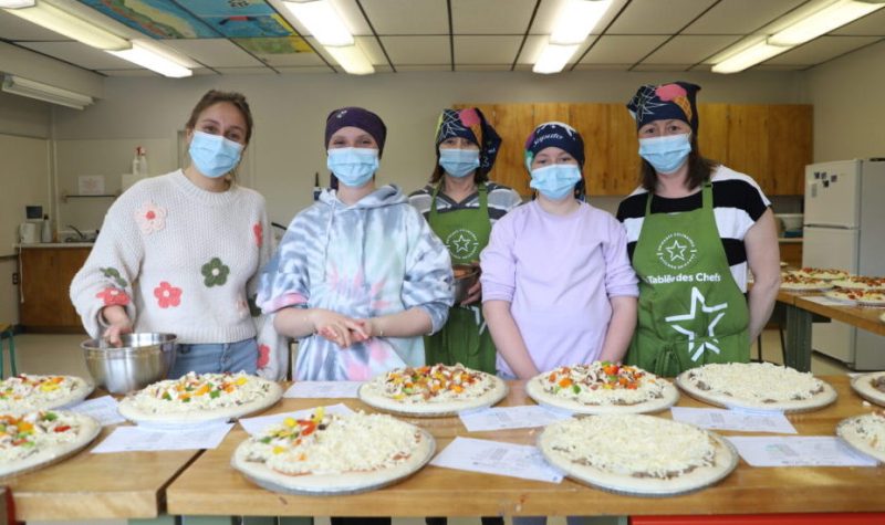 Deux élèves et trois femmes adultes portant un masque et un bandeau sur la tête posent devant des pizzas en cours de préparation.