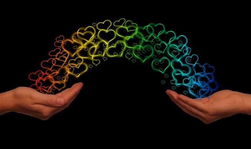 two hands reaching out to eachother. Rainbow hearts are shown making a rainbow between both hands.