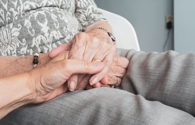 A person's hands being held by another person's hand. The main person's grey pants and white and grey shirt can also be seen.