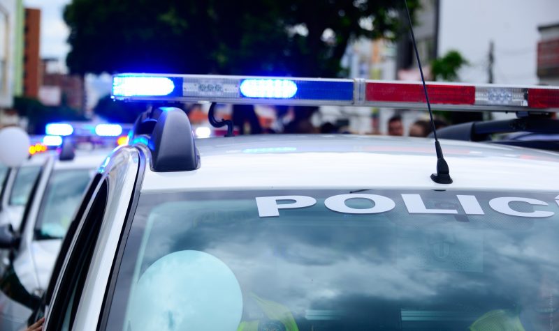 The top of a police car. Red and blue lights flashing on the roof of a police car. 2 figures are seen sitting in the car through the windshield. Police is written in white lettering on top of the windshield.