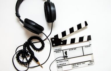 Une paire d'écouteur noir situé en haut à gauche de l'image, accompagné d'un clap cinématographique situé en dessous à droite de l'écouteur sur fond blanc.
