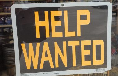 Orange lettered help wanted sign on a black background, sign is in a store window.