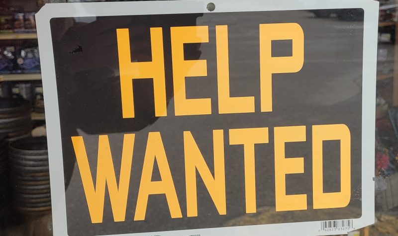 Orange lettered help wanted sign on a black background, sign is in a store window.