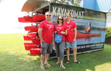 Owners/Henne Family standing in front of kayak rental rack in grass park area. It is a sunny day and they wear red shirts.