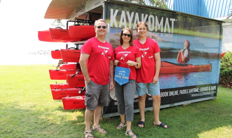 Owners/Henne Family standing in front of kayak rental rack in grass park area. It is a sunny day and they wear red shirts.