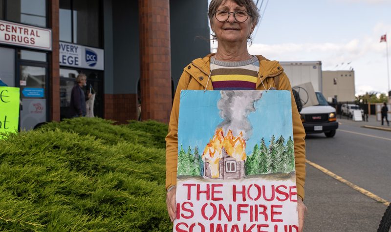 A woman stands in front of an office building wearing a sign reading the house is on fire so wake up.