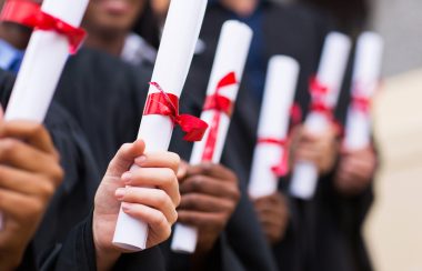 Une rangée d'étudiants tiennent leur diplôme d'une main, enroulée comme un parchemin avec un ruban rouge pour le retenir.