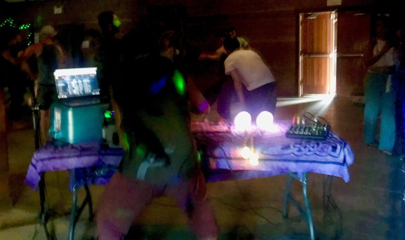 Dancers blur across a dancefloor behind a dj table.