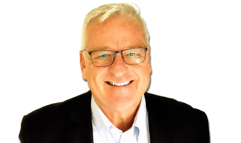 A professional head shot of Don Joyce. He is standing against a white background wearing a black suit, white dress shirt, and glasses.