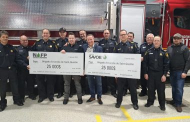 13 hommes en uniformes se tenant derrière 2 chèques format géant de 25 000 dollars respectivement. Derrière eux se trouve un camion de pompier. À l'intérieur d'une caserne de pompier