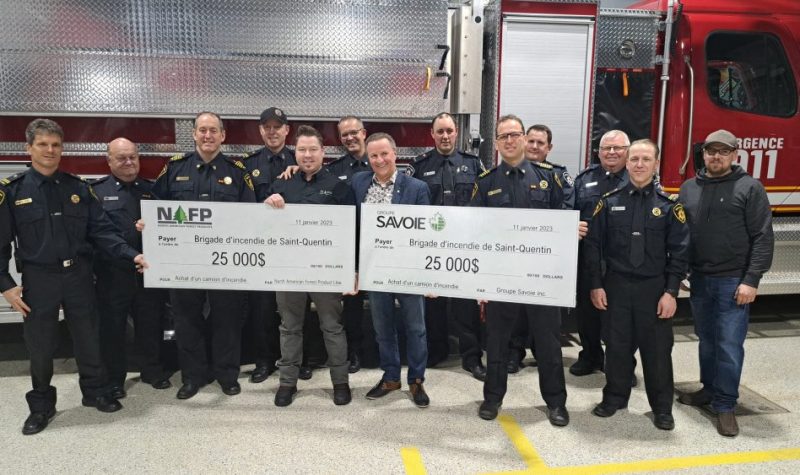 13 hommes en uniformes se tenant derrière 2 chèques format géant de 25 000 dollars respectivement. Derrière eux se trouve un camion de pompier. À l'intérieur d'une caserne de pompier