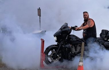 Un homme en camisole noir sur un moto noir entouré de boucane fait par le frottement de son pneu arrière sur du béton.