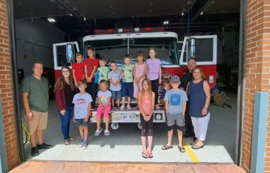 douze enfants et trois adulte devant un camion de pompier ayant les deux portes aux extrémités ouvertes, devant une entrée de caserne de pompier en plein jour