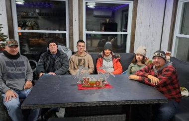 Six personnes sont assises autour d'une table noire. Derrière eux des fenêtres et un sapin de noël sur la gauche