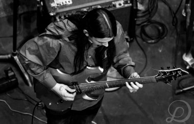 A black and white image shows a long haired musician viewed from above, playing an electric guitar.