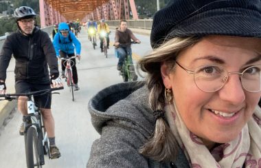 A woman taking a selfie while riding a bike across a bridge. Several other cyclists behind.