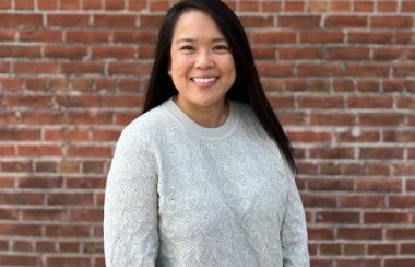 A person with long black hair is smiling while they wear a light grey sweater as they stand in front of a red brick wall.