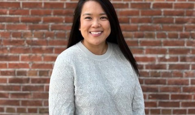A person with long black hair is smiling while they wear a light grey sweater as they stand in front of a red brick wall.