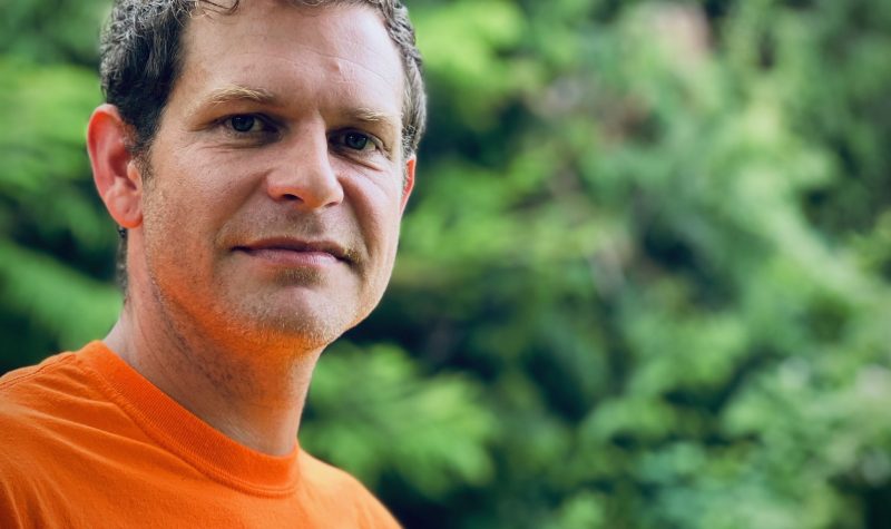 A blonde haired man in an orange shirt stands in front of a forest background.