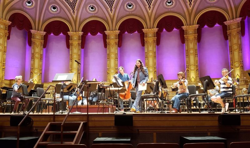 Classical musicians sit on an elevated stage with columns and a purple background.