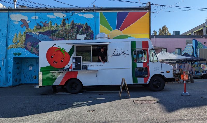 A new food truck white with a smiling tomato on one side and the name of the restaurant Lucente's is printed on the other side of the serving window. The Italian flag is painted on the door of the truck. In the background there is a mural with a colourful sun and a floating island