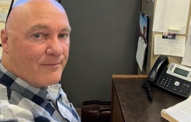 A man sitting at a desk. He is wearing a plaid shirt.