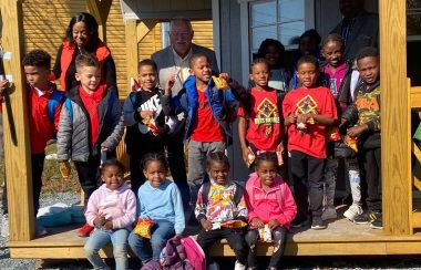 15 kids with a man and a woman, sitting in front of North Preston Society. They are smiling and looking at the camera. The children's are about 5-8 years old.