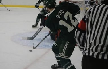 A hockey player with Boyd 12 written on his back passes the puck in front of the net.