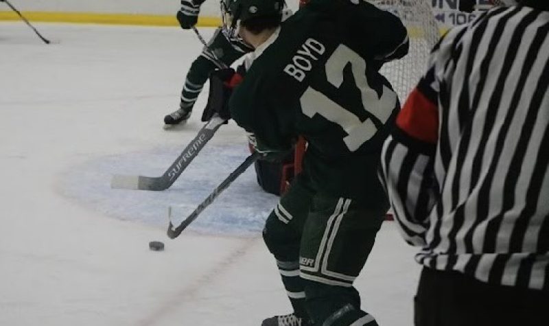A hockey player with Boyd 12 written on his back passes the puck in front of the net.