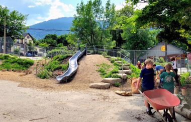 Part of the new playground. A slide goes down a dirt bank. Plants growing around. Children pushing a wheelbarrow on the side.